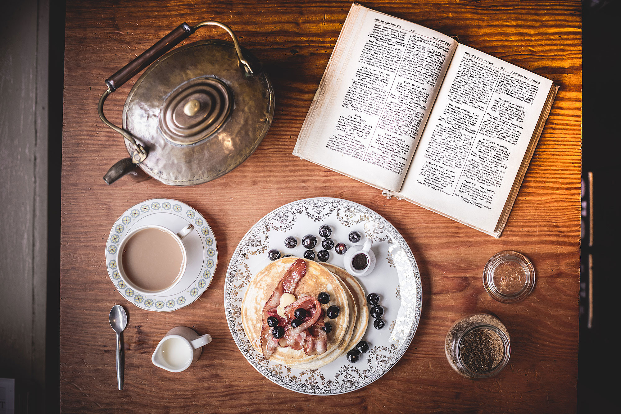 Image of an old-fashioned kettle, a cup of tea, a book and a plate of pancakes with blueberries 